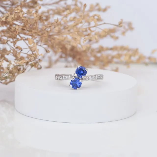 Beautiful close-up of a sapphire and diamond engagement ring on a minimalist white display with dried floral accents.